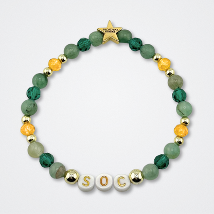 Broadway Stacks STAY GOLD bracelet inspired by The Outsiders Musical. Green and gold beads with letter beads that spell SOC. Logo star bead to remind you that YOU SHINE. Displayed on a gray background.