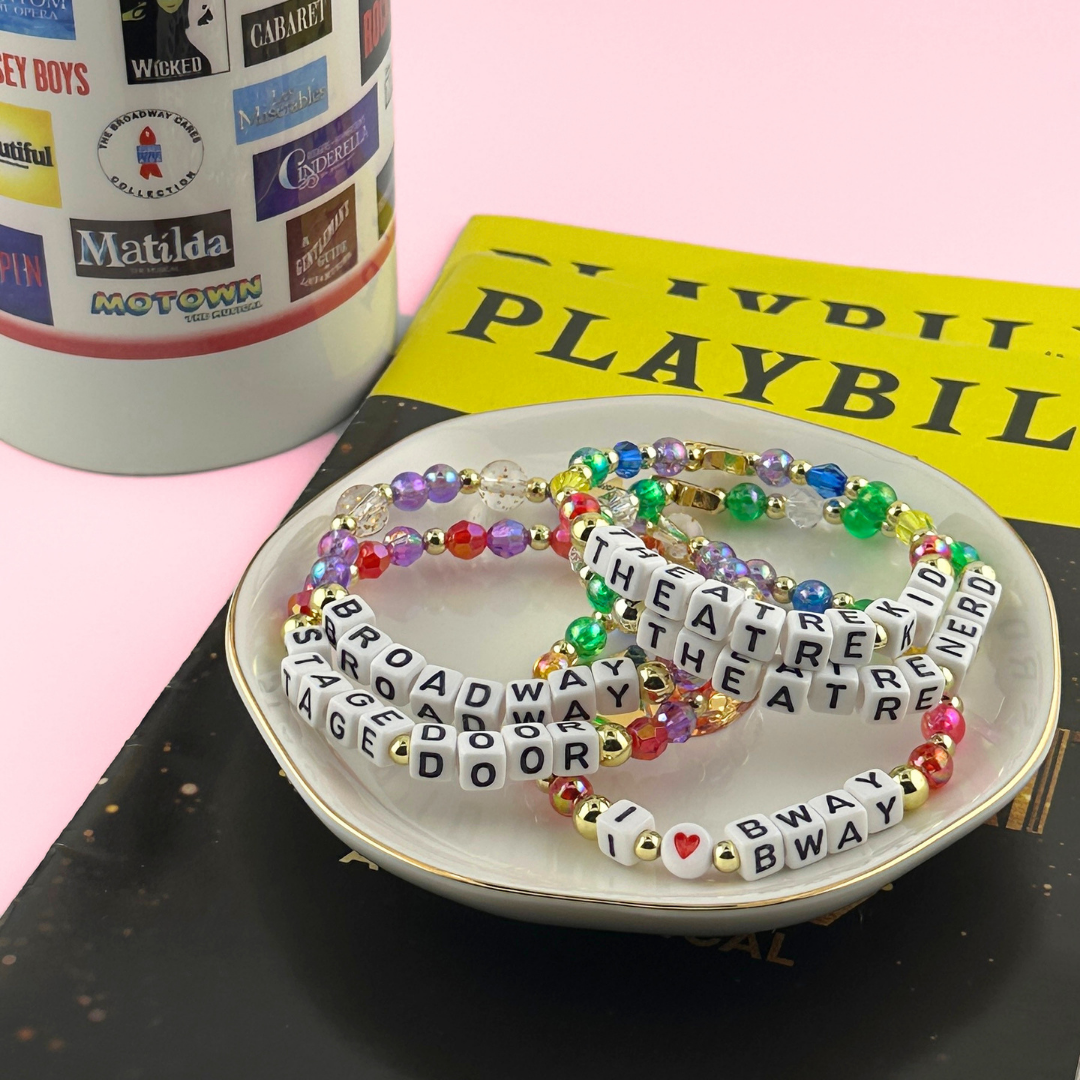 Broadway Stacks TAKE A BOW Bracelet Collection displayed in jewelry dish with playbills and theatre mug. Letter beads spell out quotes of Broadway, I love Bway, Theatre Kid, Theatre Nerd and Stage Door.
