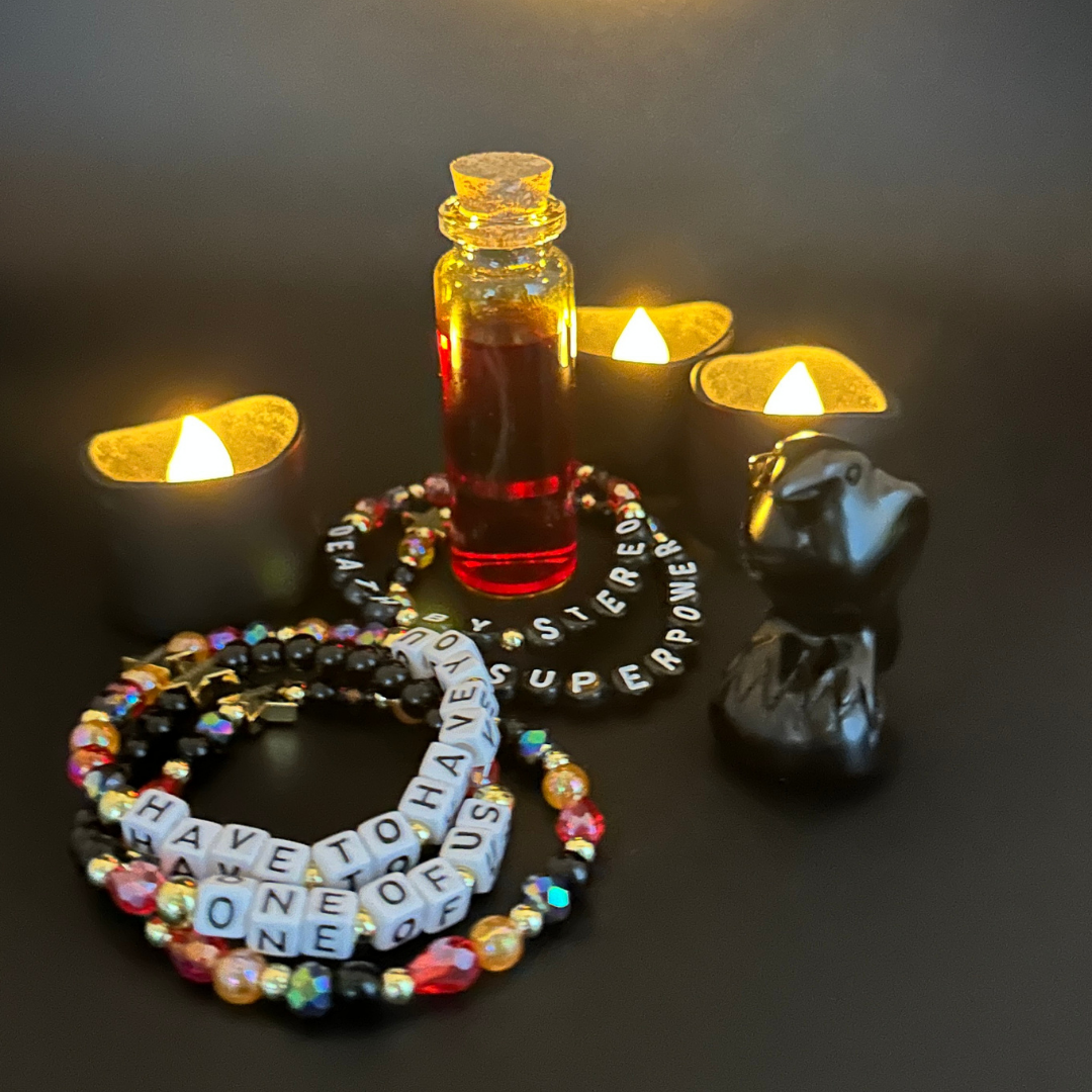 Broadway Stacks VAMPIRE Collection bead bracelets on a dark gray background. Candles lit, vial of blood and raven decor. Black, red, orange and gold colored beads with letter beads spelling out quotes. Inspired by The Lost Boys Musical.