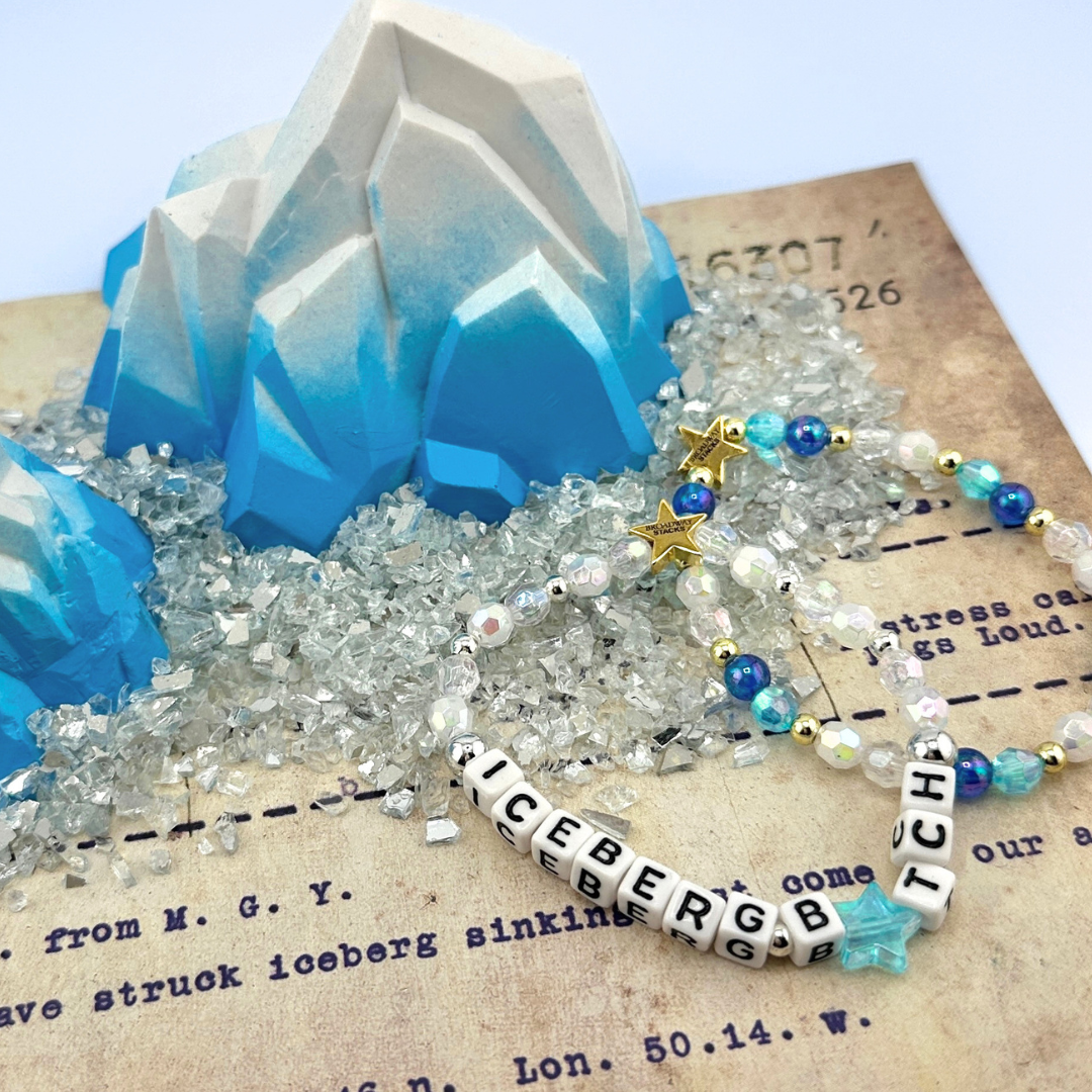 Iceberg-shapes with two beaded bracelets on an old document. Part of the Broadway Stacks UNSINKABLE Collection. Inspired by Titanique the Musical. Letter bracelet spells ICEBERG B*TCH with  blue star bead for the letter I.