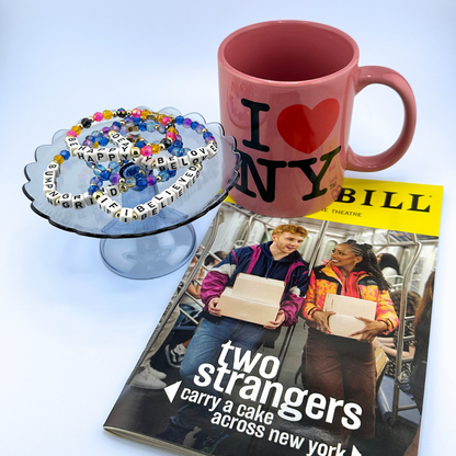 Pink mug with 'I ❤️ NY' text, cake stand with letter beads, and Playbill titled 'Two Strangers Carry a Cake Across New York' on a light blue background. Broadway Stacks CAKE Collection.