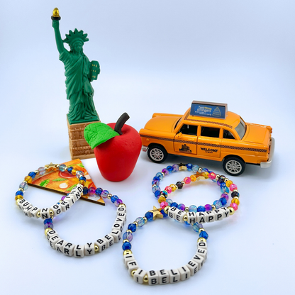 Five beaded bracelets from Broadway Stacks CAKE Collection, a toy taxi, red apple, and Statue of Liberty figurine on a light blue background.