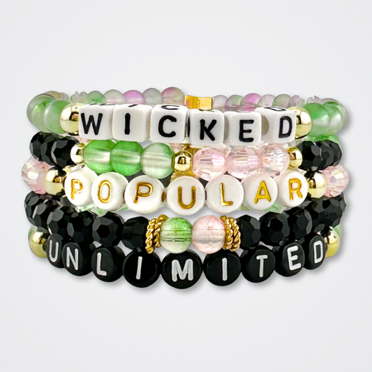 Stack of beaded bracelets with letter beads spelling out 'Wicked', 'Popular', and 'Unlimited' on a grey background. Broadway Stacks WITCH collection. Greens, pinks, black and gold colored beads. 