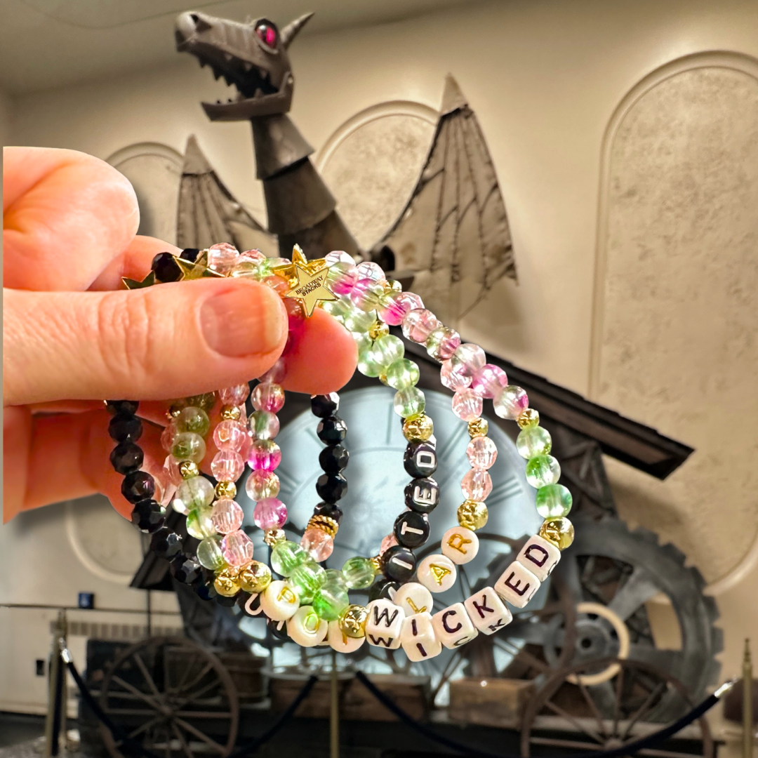Hand holding beaded bracelet stack with 'Wicked' text in front of a dragon background. Broadway Stacks WITCH collection. Greens, pinks, black and gold colored beads. Gold star logo bead to remind you that You Shine. 