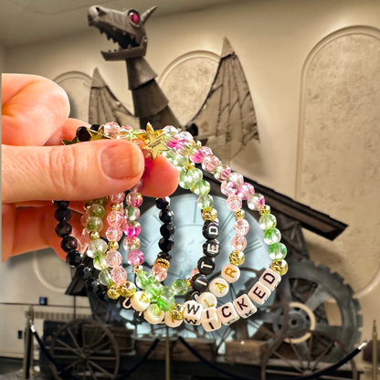 Hand holding beaded bracelet stack with 'Wicked' text in front of a dragon background. Broadway Stacks WITCH collection. Greens, pinks, black and gold colored beads. Gold star logo bead to remind you that You Shine. 