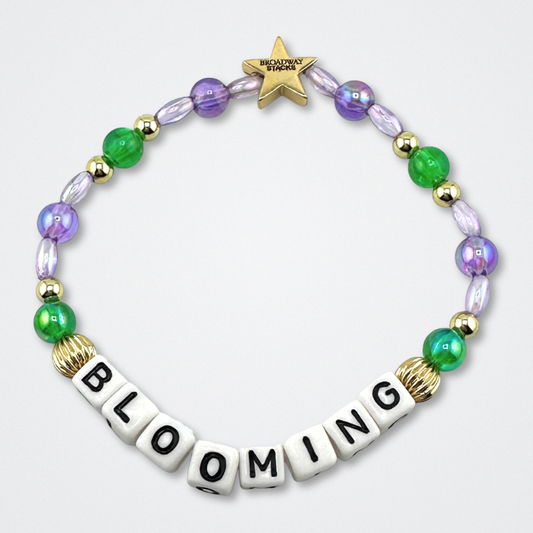 Broadway Stacks GARDEN bracelet with green, purple and gold color beads. Letters say BLOOMING. Displayed on a gray background.