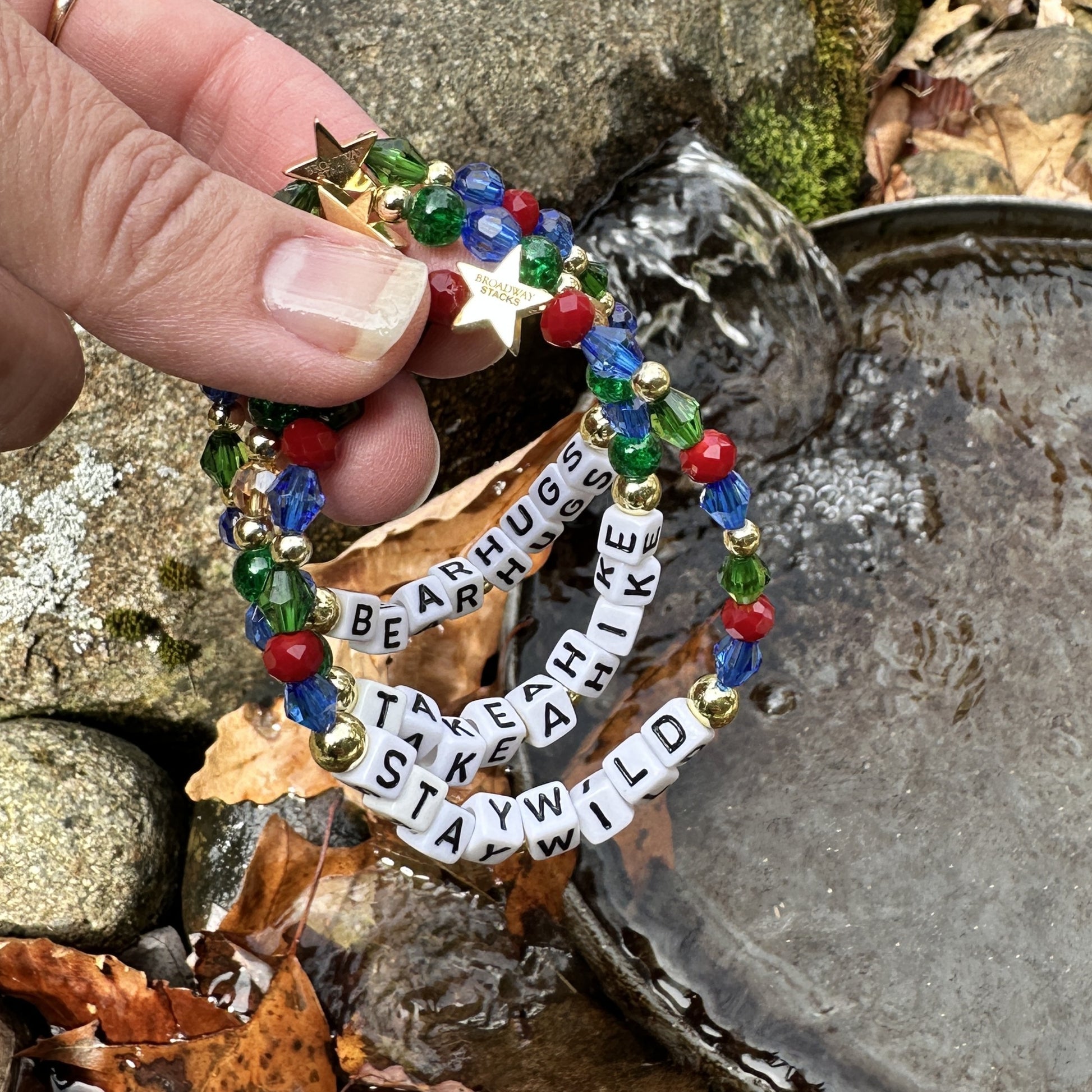 Broadway Stacks MOUNTAIN Collection bead bracelets. Green, blue, dark red and gold color jewelry. Hand holding bracelets over rocks and water.