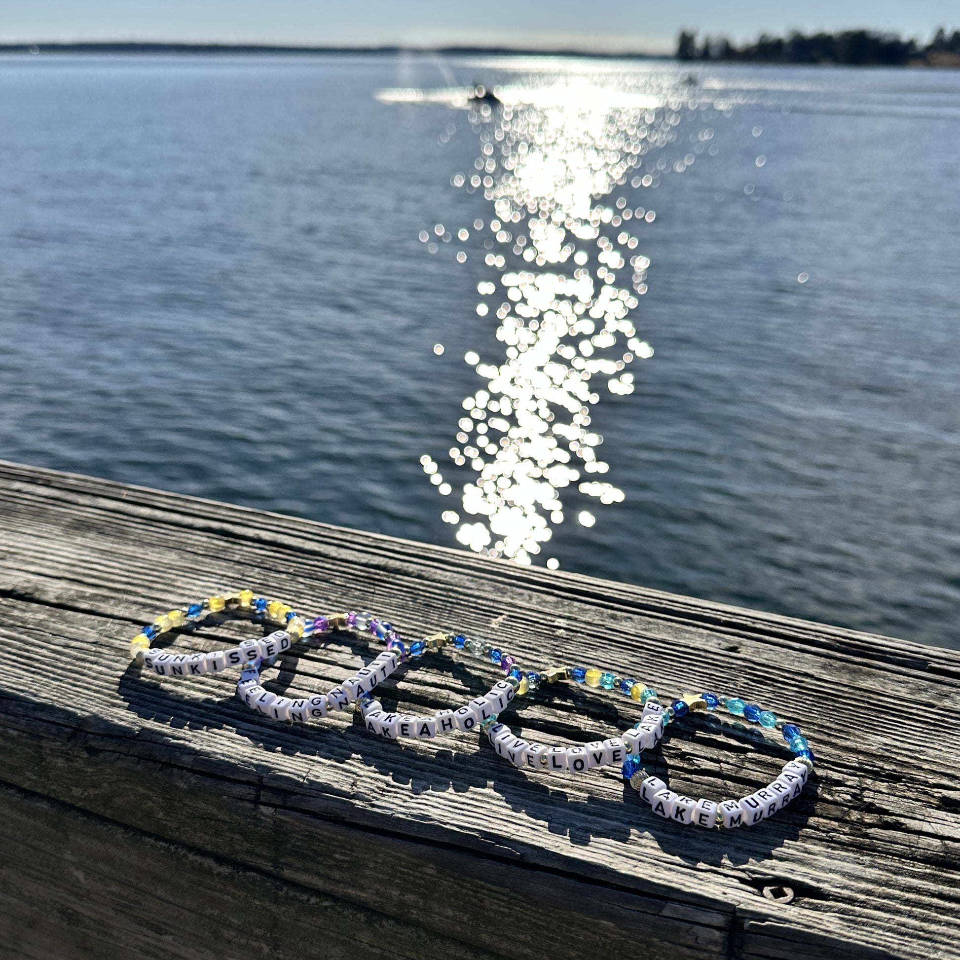 Broadway Stacks LAKE LIFE bracelet collection. Blues, yellow, purple and gold color bead bracelets displayed on dock railing. Sun shining on water in background.