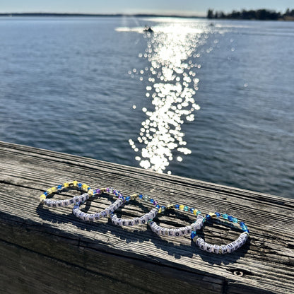 Broadway Stacks LAKE LIFE bracelet collection. Blues, yellow, purple and gold color bead bracelets displayed on dock railing. Sun shining on water in background.