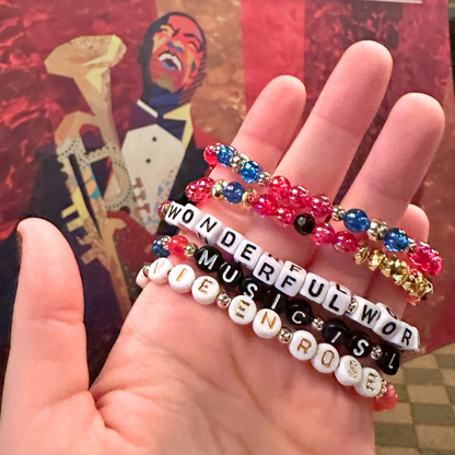Broadway Stacks JAZZY Collection. 5 stretch bracelets included in Stack. Red, blue, black and gold colored beads. Letter beads that spell quotes. Gold star logo bead to remind you that you shine. Wonderful World musical and Louis Armstrong fans.